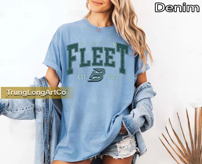 May include: Light blue t-shirt with the text "BOSTON FLEET" in dark green block letters. The shirt also features the text "EST 2023" and a stylized "B" logo. The word "Denim" is in the upper right corner.