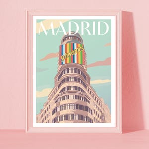 May include: A framed art print featuring a stylized illustration of a building in Madrid. The building has a curved facade and a colorful Schweppes sign. The word "MADRID" is printed at the top against a light blue sky.