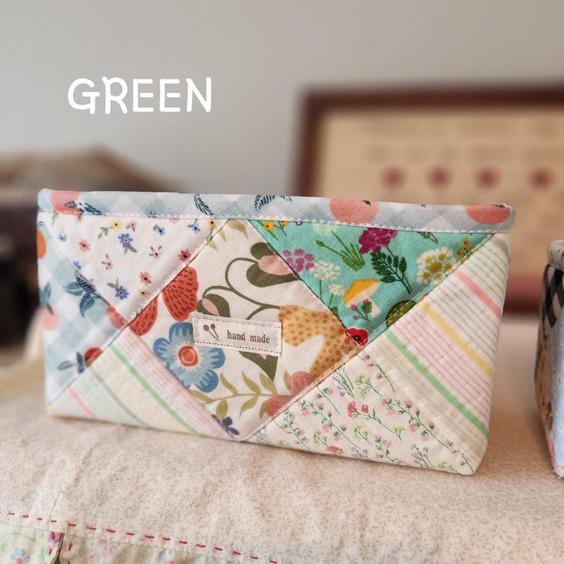 Pode incluir: Uma bolsa acolchoada com um design de patchwork com padr&otilde;es florais e geom&eacute;tricos em tons de azul, verde, rosa e branco. O texto "GREEN" est&aacute; no topo e uma pequena etiqueta diz "hand made".