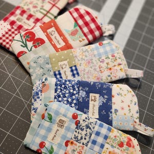 May include: Four rectangular fabric pouches featuring patchwork designs in red, blue, and green floral, gingham, and fruit patterns. Each pouch has a small fabric loop at the top and a white label with the text Hand Made and a sewing icon.