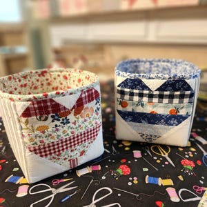 May include: Two fabric storage baskets with heart-shaped patchwork designs. One basket features a red and white floral and gingham pattern, while the other has a blue and white floral and gingham pattern. The baskets sit on a black sewing-themed fabric.