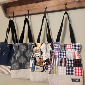 May include: Four tote bags with black straps hang from hooks. The bags feature various patterns: a navy floral, a gray tree design, a dog print, and a patchwork of plaid fabrics. Each bag has a neutral-colored bottom.