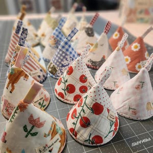 May include: A collection of cone-shaped fabric tea cozies featuring various patterns including red tomatoes, floral prints, gingham checks, and animal motifs. Each cozy has a small fabric loop at the top and a small tag labeled Hand Made.