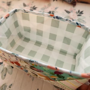 Pode incluir: Uma cesta retangular de tecido com um padr&atilde;o floral e xadrez. O interior apresenta um design xadrez verde claro e branco. O exterior tem um padr&atilde;o floral colorido com toques de azul, rosa e laranja. A cesta &eacute; provavelmente feita &agrave; m&atilde;o e adequada para armazenamento.