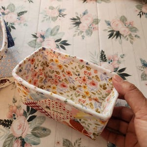 Pode incluir: Uma cesta retangular de tecido com um padr&atilde;o floral no interior e um design de patchwork no exterior. A cesta &eacute; segurada por uma m&atilde;o, mostrando a qualidade artesanal. O interior apresenta uma impress&atilde;o floral colorida.