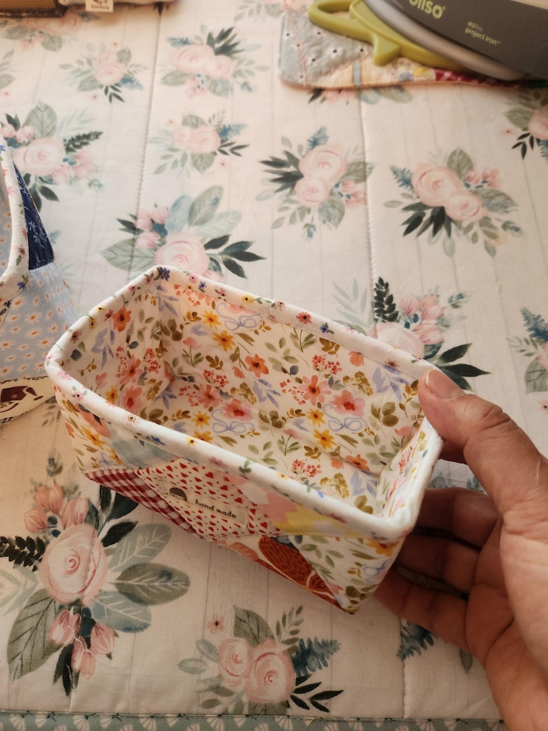 Pode incluir: Uma cesta retangular de tecido com um padr&atilde;o floral no interior e um design de patchwork no exterior. A cesta &eacute; segurada por uma m&atilde;o, mostrando a qualidade artesanal. O interior apresenta uma impress&atilde;o floral colorida.