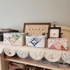 Pode incluir: Tr&ecirc;s cestos de armazenamento de tecido feitos &agrave; m&atilde;o em tamanhos variados. Os cestos s&atilde;o feitos de tecido de patchwork em uma variedade de cores e padr&otilde;es, incluindo designs florais, xadrez e listrados. Os cestos s&atilde;o exibidos em uma prateleira com uma borda decorativa ondulada.