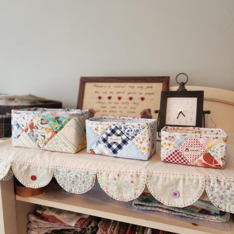 Pode incluir: Tr&ecirc;s cestos de armazenamento de tecido feitos &agrave; m&atilde;o em tamanhos variados. Os cestos s&atilde;o feitos de tecido de patchwork em uma variedade de cores e padr&otilde;es, incluindo designs florais, xadrez e listrados. Os cestos s&atilde;o exibidos em uma prateleira com uma borda decorativa ondulada.