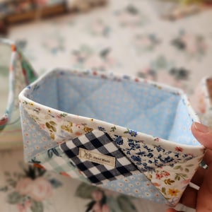 Pode incluir: Cesto de armazenamento de tecido feito &agrave; m&atilde;o com um design de patchwork. O cesto apresenta um interior azul claro com um padr&atilde;o acolchoado. O exterior &eacute; uma mistura de tecidos florais, xadrez e de bolinhas. Uma pequena etiqueta diz "Hand Made".