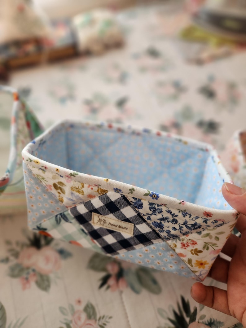 Pode incluir: Cesto de armazenamento de tecido feito &agrave; m&atilde;o com um design de patchwork. O cesto apresenta um interior azul claro com um padr&atilde;o acolchoado. O exterior &eacute; uma mistura de tecidos florais, xadrez e de bolinhas. Uma pequena etiqueta diz "Hand Made".