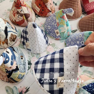 May include: A collection of quilted pot holders in various patterns, including floral, gingham, and animal prints. The pot holders are in shades of blue, white, red, and beige. The text "YOU'RE INVITED TO Julie's FarmHouse" is visible on one of the pot holders.