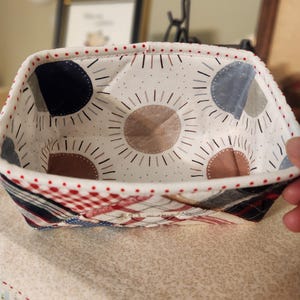 Pode incluir: Uma cesta de tecido feita &agrave; m&atilde;o com exterior e interior estampados. O interior apresenta um fundo branco com desenhos de raios de sol castanhos, azuis e cinzentos. O exterior tem um design de patchwork em vermelho, branco e azul.