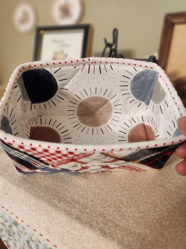 Pode incluir: Uma cesta de tecido feita &agrave; m&atilde;o com exterior e interior estampados. O interior apresenta um fundo branco com desenhos de raios de sol castanhos, azuis e cinzentos. O exterior tem um design de patchwork em vermelho, branco e azul.