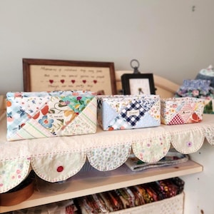 Pode incluir: Tr&ecirc;s caixas de armazenamento de tecido feitas &agrave; m&atilde;o em v&aacute;rios tamanhos, com designs de patchwork com padr&otilde;es florais e geom&eacute;tricos. As caixas s&atilde;o em tons de azul, rosa e branco, com uma prateleira com borda ondulada em primeiro plano. Uma obra de arte emoldurada e um pequeno rel&oacute;gio est&atilde;o no fundo.