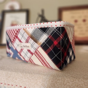 Pode incluir: Uma cesta retangular de tecido com um design de patchwork. A cesta apresenta uma mistura de padr&otilde;es xadrez e listrados vermelhos, brancos, azuis e pretos. A borda superior &eacute; forrada com tecido branco e bolinhas vermelhas. Uma pequena etiqueta diz "Hand Made".