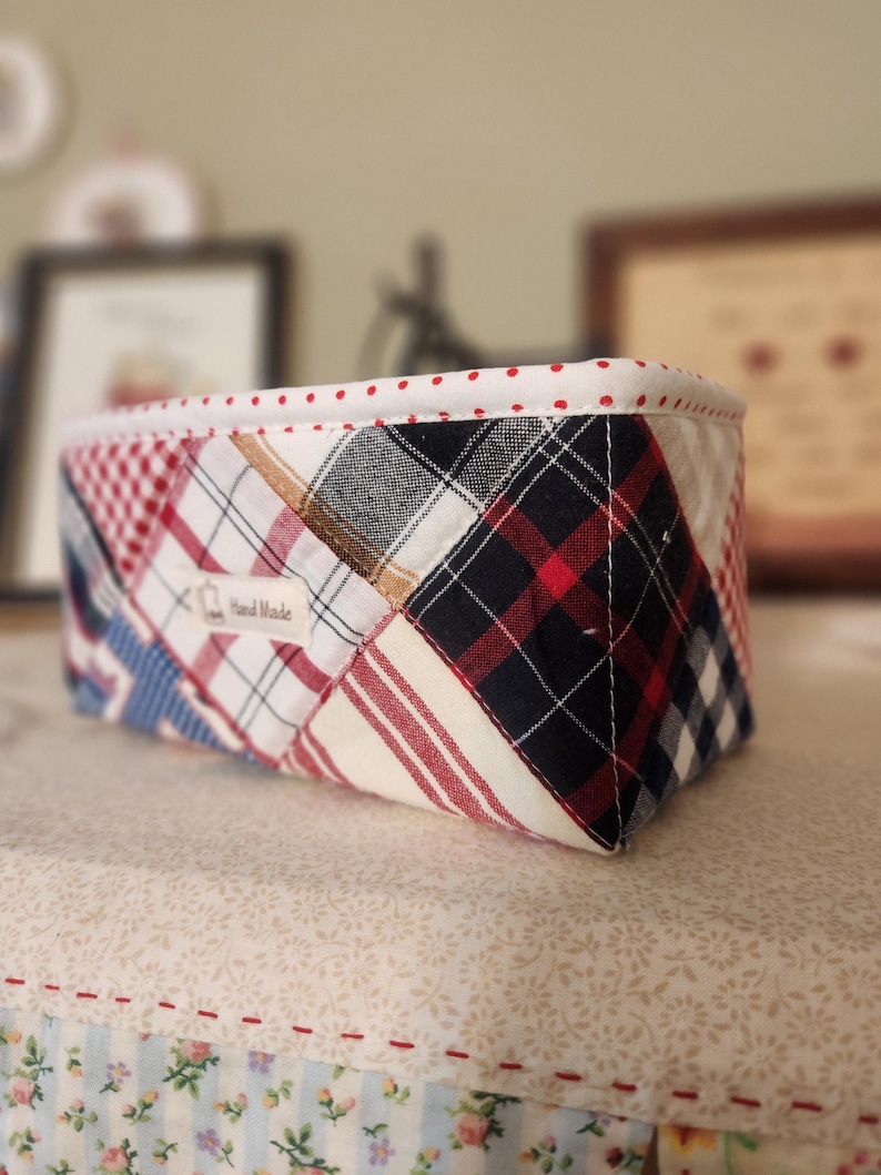 Pode incluir: Uma cesta retangular de tecido com um design de patchwork. A cesta apresenta uma mistura de padr&otilde;es xadrez e listrados vermelhos, brancos, azuis e pretos. A borda superior &eacute; forrada com tecido branco e bolinhas vermelhas. Uma pequena etiqueta diz "Hand Made".