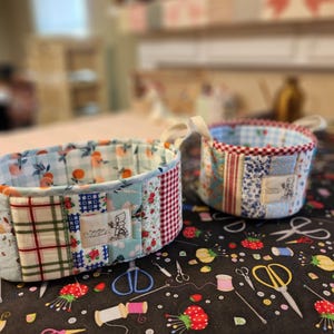 May include: Two small, handmade fabric baskets with patchwork designs. One basket features a light blue floral pattern, while the other has a red and blue patchwork design. Both have cream-colored handles. The baskets sit on a black surface with sewing-themed illustrations.
