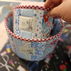 May include: Two round fabric baskets featuring a patchwork design of blue, white, and red floral patterns. Each basket has a red and white gingham trim and a rectangular fabric label on the front depicting a child in a hooded cloak.