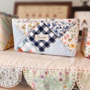 May include: A small, rectangular fabric basket with a patchwork design. The basket features a mix of floral, gingham, and solid blue fabrics. A small label reads "Hand Made". The basket is sitting on a shelf with a scalloped edge.