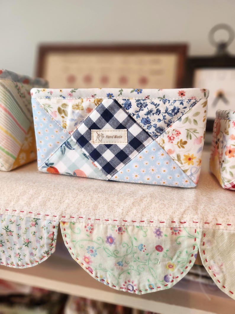 Pode incluir: Uma pequena cesta de tecido feita &agrave; m&atilde;o com um design de patchwork. A cesta apresenta uma mistura de tecidos florais, xadrez e azuis lisos. Uma pequena etiqueta diz "Hand Made". A cesta est&aacute; em uma prateleira com borda ondulada.