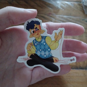 Puede incluir: Una pegatina con un personaje de dibujos animados con orejas puntiagudas, que lleva una camisa amarilla y un chaleco azul con rombos. El personaje está sentado, con hilo rojo enrollado alrededor de sus extremidades y una bobina de hilo. El personaje tiene un ojo morado.