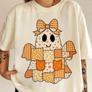 May include: Cream-coloured t-shirt with a patchwork ghost design. The ghost has a bow, eyes, and a smiling mouth. The patchwork includes orange gingham, floral, and solid orange patterns. The shirt is a relaxed fit.