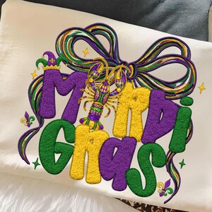 May include: Cream-colored t-shirt with a colorful Mardi Gras design. The design features the words "Mardi Gras" in green, yellow, and purple, with a bow and a crawfish. The shirt is folded on a white surface.