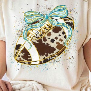 May include: Cream-colored t-shirt featuring a football design. The football has a brown and white cowhide pattern, accented with gold trim and a light blue bow. The design is embellished with blue and gold paint splatters.