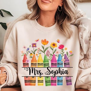 Puede incluir: Sudadera crema con un diseño de crayones coloridos y flores. El diseño incluye crayones rojos, naranjas, amarillos, verdes, rosas, azules y morados, con el texto "Mrs. Sophia".