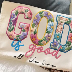 Op de afbeelding: Cr&egrave;mekleurig T-shirt met de geborduurde tekst "GOD is good all the time." De letters zijn gevuld met kleurrijke bloemmotieven in roze, paars, blauw en geel. Het shirt ligt op een witte, zachte ondergrond.