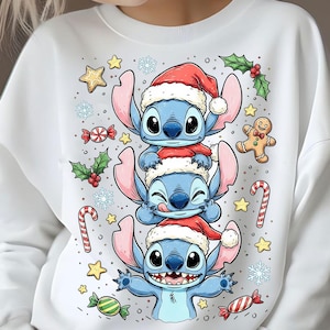 May include: White sweatshirt featuring a stacked design of three blue cartoon characters wearing Santa hats. The design includes candy canes, stars, snowflakes, and gingerbread men, creating a festive holiday theme.