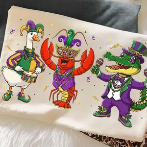 May include: A cream-colored t-shirt featuring cartoon characters dressed in Mardi Gras attire. A goose plays a drum, a lobster wears a mask, and an alligator holds maracas. The characters are in purple, green, and gold.