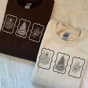 May include: Two sweatshirts, one brown and one beige, each featuring a decorative design. The design includes a mistletoe sprig, a Christmas tree, and a poinsettia, all within ornate frames. The sweatshirts are folded.