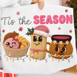 May include: White pillow with the text "Tis the SEASON" in black and pink. The pillow features embroidered images of a basket of pastries, a tamale with a Santa hat, and a mug with a floral crown. A string of lights is at the bottom.