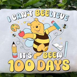 May include: White sweatshirt featuring a cartoon Winnie the Pooh character in a bee costume, holding an apple and pencil. Text reads "I CAN'T BEE-LIEVE IT'S BEEN 100 DAYS" with school-themed graphics.