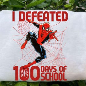 Puede incluir: Camiseta blanca con un gráfico de Spider-Man en una pose dinámica, con detalles de traje rojo y negro. El texto "I Defeated 100 Days of School" está impreso en rojo, junto con un diseño de telaraña. Una camiseta divertida y temática.