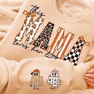 May include: Beige sweatshirt with the text "this MAMA loves her littles". The word "MAMA" is designed with pumpkin, leopard print, and striped patterns. Two ghost illustrations are on the sleeve, one with a baseball cap and the other with a bow.