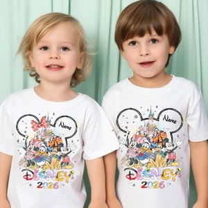May include: Two white t-shirts with a colorful Disney graphic featuring Mickey Mouse, Minnie Mouse, Donald Duck, and other characters. The shirts have a Mickey Mouse ear outline with the word "Name" and the year "2026".