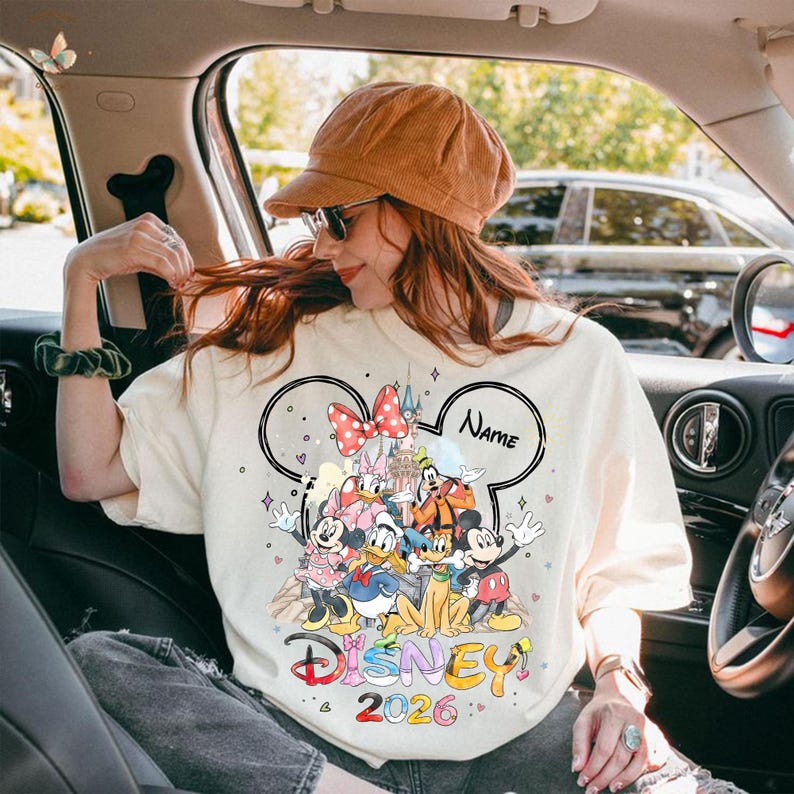 May include: Cream-colored Disney-themed sweatshirt featuring a large graphic of Disney characters, a castle, and a red polka-dot bow. The word "Disney" is written in colorful letters, with "2026" below. The design is framed by a Mickey Mouse head shape, with a space for a name.