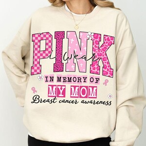 K&ouml;nnte beinhalten: Cremefarbenes Sweatshirt mit den Worten "PINK I wear" in Rosa, mit verschiedenen Mustern und einem Blumenakzent. Darunter steht "IN MEMORY OF MY MOM" und "Breast cancer awareness" in Rosa und Schwarz.