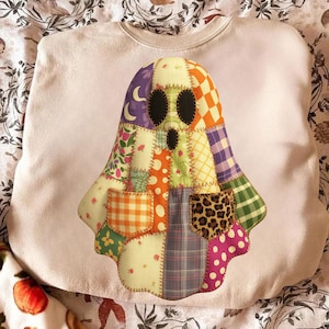 May include: A pale pink sweatshirt with a patchwork ghost design. The ghost is constructed from diverse fabric patterns, including floral, plaid, and leopard print. The ghost has black oval eyes and a small mouth.