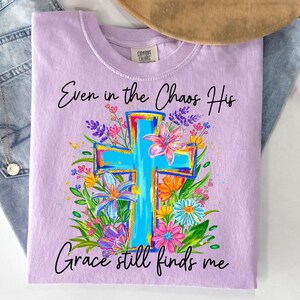 May include: A light purple t-shirt with the text "Even in the Chaos His Grace still finds me." The shirt features a blue cross design surrounded by colorful flowers in shades of pink, yellow, and purple.