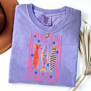 Pode incluir: Uma camiseta Comfort Colors lavanda com um design de peixes coloridos. O gr&aacute;fico inclui tr&ecirc;s peixes com padr&otilde;es diferentes, listras verticais rosa e estrelas azuis. Um colar de ouro repousa sobre a camiseta.