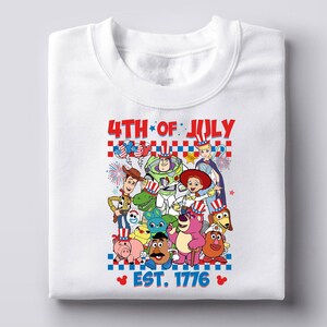 May include: White t-shirt with a graphic of the Toy Story characters wearing red, white, and blue patriotic hats. The text "4th of July Est. 1776" is printed below the characters.