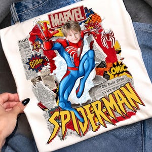May include: A white t-shirt featuring a graphic print of a child's face superimposed on a Spider-Man character body. The design includes comic book style elements, newspaper clippings, the text MARVEL, BAM, COMIC, and SPIDER-MAN in yellow.