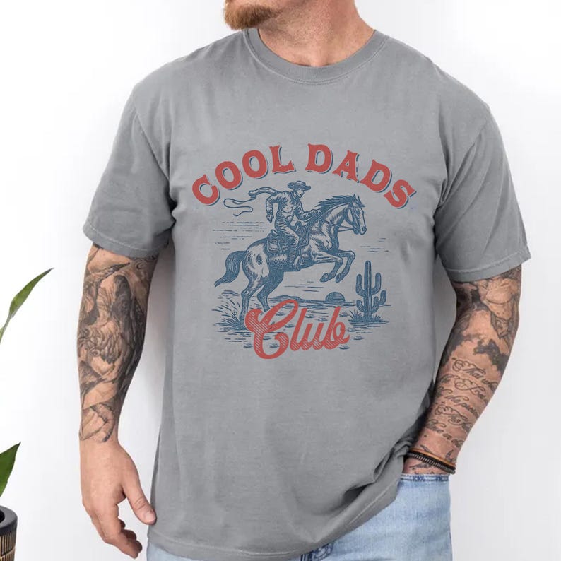 May include: Gray t-shirt with a graphic design featuring a cowboy riding a horse, a cactus, and the words "COOL DADS Club" in red. The shirt is a solid color and has short sleeves.