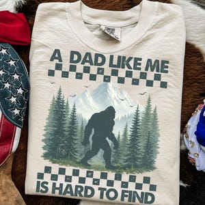 May include: Cream-colored t-shirt with the text "A DAD LIKE ME IS HARD TO FIND." The shirt features a graphic of a Bigfoot walking through a forest with mountains in the background. The design includes a checkered pattern.
