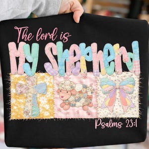 May include: Black sweatshirt with the text "The lord is MY Shepherd" in pastel colors. Below the text are three patchwork designs: a cross, a sheep, and a bow. The text "Psalms 23:1" is at the bottom.