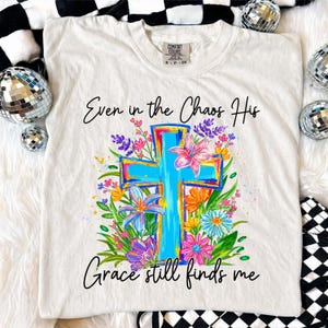 May include: Cream-colored t-shirt with a vibrant, floral cross design. The cross is outlined in bright colors, with a blue center and surrounded by colorful flowers. The text reads "Even in the Chaos His Grace still finds me."