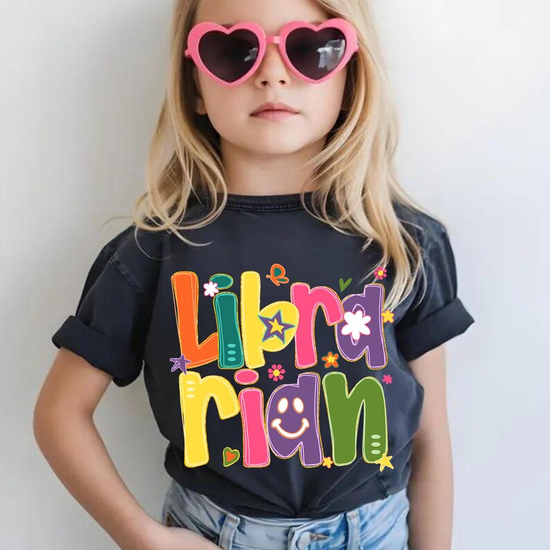 May include: A dark gray t-shirt with the word "Librarian" in colorful, playful letters. The design includes flowers, stars, and a smiling face. The shirt is worn by a young person wearing pink heart-shaped sunglasses.